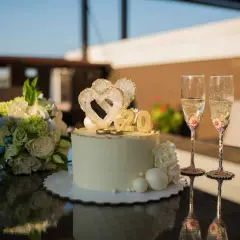Torta per 20 anni di matrimonio con cuori, anelli e numero 20 dorato. Due flute di champagne decorate e un bouquet di fiori completano la scena.