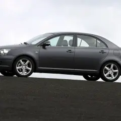 Szary sedan Toyota Avensis 1.8 benzyna stoi na ciemnej nawierzchni, z niebieskim niebem w tle.