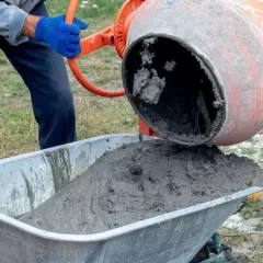 Béton frais coulant d'une bétonnière orange dans une brouette. Un ouvrier en gants bleus manipule la manivelle pour déverser le mortier béton.