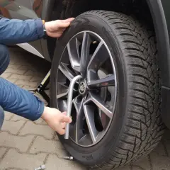 Osoba w niebieskiej kurtce i jeansach dokonuje wymiany koła w samochodzie, używając podnośnika i klucza.