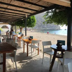 Mesas y sillas en el exterior del restaurante Agua Amarga, con la playa y el mar de fondo.