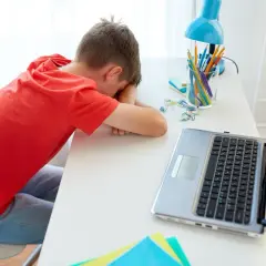 Chłopiec w czerwonej koszulce śpi na biurku obok laptopa i przyborów szkolnych.