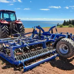 Czerwony traktor z niebieskim agregatem uprawowym na polu, gotowy do siewu. W tle błękitne niebo i morze.