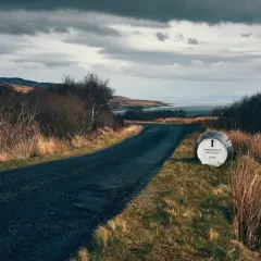 Route sinueuse menant à la distillerie Bunnahabhain, avec un fût indiquant la direction. Paysage écossais sauvage sous un ciel menaçant.