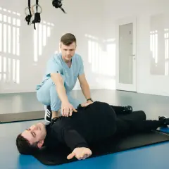 Un kinésithérapeute aide un patient allongé sur un tapis à réaliser un mouvement pour le diagnostic du syndrome du piriforme.