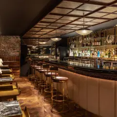Interni eleganti di un club a Londra, con un lungo bancone bar illuminato, sgabelli dorati e un murale tropicale.