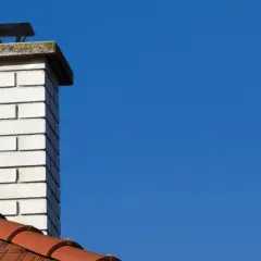 Biały, ceglany komin spalinowy z metalową nasadką na tle błękitnego nieba. Obok fragment dachu pokryty czerwonymi dachówkami.