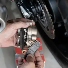 Mani che effettuano il cambio pastiglie freni di una moto, mostrando le vecchie pastiglie usurate e il nuovo set pronto per l'installazione.