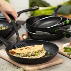 Eine Hand wendet mit einem Pfannenwender einen gefüllten Pfannkuchen in einer beschichteten Pfanne. Mehrere Pfannen und Zutaten wie Pilze und Eier sind im Hintergrund zu sehen.
