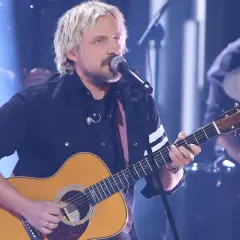 Artysta z blond włosami gra na gitarze akustycznej, śpiewając do mikrofonu. Juz teraz wiem ze dni tekst.