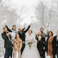 Para ślub zimą jak się ubrać? Para młoda i goście w zimowym lesie rzucają śnieżkami. Panna młoda w białym futerku, pan młody w eleganckim płaszczu.