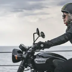 Mujer rubia en moto Triumph negra, con casco y chaqueta de cuero, disfrutando del paisaje. Las motos Triumph se fabrican con pasión.