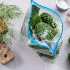 Przygotowanie ogórków małosolnych bez wody. W przezroczystej torbie zamykanej na zamek błyskawiczny znajdują się świeże ogórki, koperek i czosnek, posypane solą.
