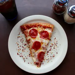 Kawałek pizzy pepperoni na talerzu, obok słoiczki z przyprawami i napój. Zastanawiasz się, ile kosztuje pizza we Włoszech?