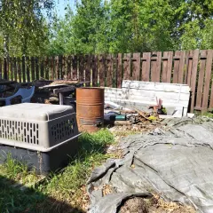 Zarośnięty ogród z rupieciami, beczką i klatką dla psa. Wygląda jak podwórko uciążliwego sąsiada na wsi.