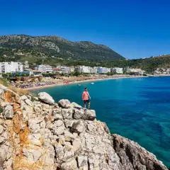 Mann steht auf Felsen mit Blick auf eine Bucht mit Strand und Hotels. Perfekt, um zu wissen, wo in Albanien Urlaub machen!