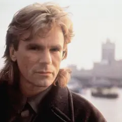 MacGyver obsada: Richard Dean Anderson jako MacGyver, z charakterystyczną fryzurą i w płaszczu, na tle mglistego miasta.