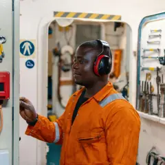 Técnico con auriculares rojos ajusta un panel de control. Herramientas cuelgan en la pared. El nivel de ruido podría superar la escala db.