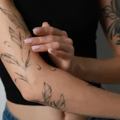 Delikatny tatuaż z liśćmi na ramieniu, jak druga skóra po tatuazu. Dłoń z zadbanymi paznokciami dotyka skóry.