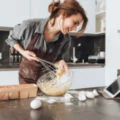 Uśmiechnięta kobieta w fartuchu miesza masę w misce, sprawdzając przepis na tablecie. Na blacie leżą jajka i mąka, jakby właśnie przygotowywała ciasto z erytrytolem.