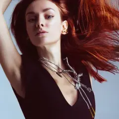 Mujer con cabello cobrizo vibrante, moviéndose con gracia. Luce un collar plateado llamativo y un pendiente dorado.