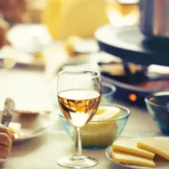 Un verre de vin blanc sec ou moelleux accompagne une raclette conviviale. Des morceaux de fromage et des cornichons sont prêts à être dégustés.
