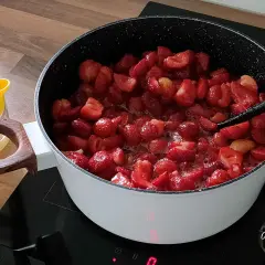 Duży garnek z gotującymi się truskawkami na kuchence indukcyjnej. Obok żółta wyciskarka do cytrusów.
