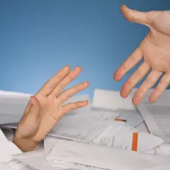 Dłoń wyciągnięta z papierów, druga dłoń oferuje pomoc. Symbolizuje trudności i potrzebę wsparcia w procesie restrukturyzacji.