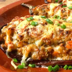 Berenjenas rellenas gratinadas con queso derretido y cebolleta picada, servidas en una cazuela de barro.