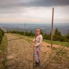 Uśmiechnięta turystka z kijkami na kamienistej ścieżce. Widok na góry i doliny. Czas wejścia na Śnieżkę zależy od kondycji i wybranej trasy.