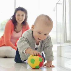 Dziecko raczkuje, ćwicząc motorykę dużą co to jest, sięgając po kolorową piłkę. Uśmiechnięta mama obserwuje.