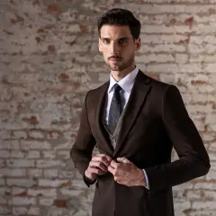 Elegancki mężczyzna w brązowej marynarce, kamizelce, białej koszuli i wzorzystym krawacie zapina marynarkę.