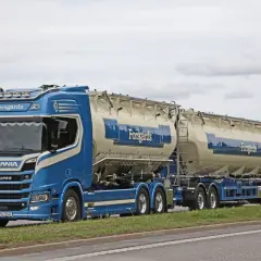 Ein blauer Scania-Sattelzug mit zwei Siloanhängern, beladen für Forsgårds, fährt auf einer schwedischen Straße. Das Tempolimit in Schweden ist zu beachten.