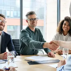 Uśmiechnięci ludzie przy stole, jeden z nich ściska dłoń, omawiając, jak udokumentować umowę zlecenie do emerytury.