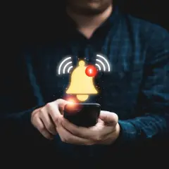 Dłoń trzymająca smartfon, nad którym unosi się złoty dzwonek z powiadomieniem.