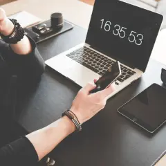 Osoba trzyma iPhone'a, obok leży tablet i laptop z widocznym zegarem.