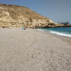 Arena y guijarros en la playa de las salinicas, con acantilados dorados y mar azul. Una sombrilla azul protege a unos bañistas.