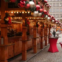 Drewniane stoiska ozdobione bombkami i lampkami tworzą świąteczną atmosferę na jarmarku we Wrocławiu. W tle widać ludzi i budynki.