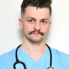 Młody rezydent w niebieskiej bluzie medycznej, ze stetoskopem na szyi i charakterystycznymi wąsami.