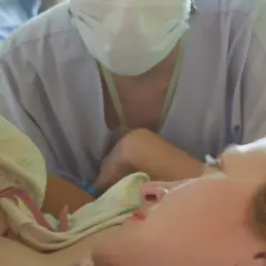 Noworodek przytulony do mamy, otoczony pieluchą. W tle widać personel medyczny w maseczkach, wspierający poród lotosowy.