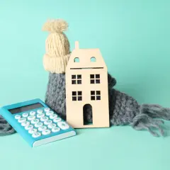 Casa de madera abrigada con bufanda y gorro, junto a calculadora. Simboliza el cálculo del coeficiente de rendimiento energético para el hogar.