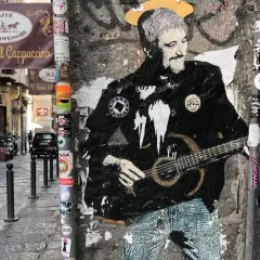 Graffiti di Pino Daniele con la chitarra, un omaggio a Napoli. I testi "Resta... resta cu'mmè" risuonano tra i vicoli.