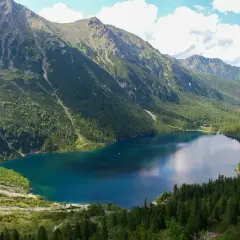 Widok na turkusowe Morskie Oko otoczone stromymi, zalesionymi górami. W oddali widać niewielki budynek.