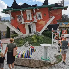 Ein rotes Haus steht auf dem Kopf, die Treppe führt ins Nichts. Eine Familie bestaunt das Spektakel, bunte Skulpturen und ein Dino-Land im Hintergrund.