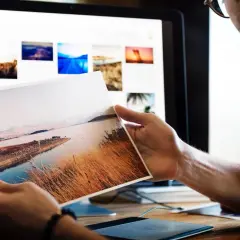 Fotograf przegląda wydrukowane zdjęcie krajobrazu, szukając inspiracji do nowych zleceń. Na ekranie komputera widać galerię jego prac.