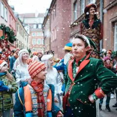 Dawid w stroju elfa prowadzi świąteczny korowód ulicą pełną ludzi i ozdób. Dzieci z uśmiechem patrzą na magię jarmarku.