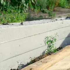 Betonowy fundament pod ogrodzenie, z widocznymi deskami szalunkowymi i zielenią.