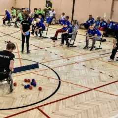 Zawodnicy grają w boccię, sportową grę precyzyjną. Na boisku widać kolorowe kule i zawodnika na wózku inwalidzkim.