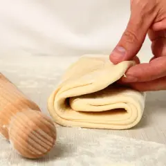 Mani che piegano pasta sfoglia, pronta per diventare pasta fatta in casa. Un mattarello è accanto, pronto per il prossimo passaggio.