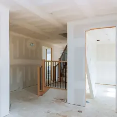 Interno di una casa in costruzione con pareti in cartongesso grezzo, una scala in legno e metallo e un'apertura verso un'altra stanza.